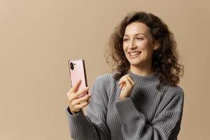 Friendly curly beautiful female in gray casual sweater doing video call with phone posing isolated on over beige pastel background. Social media, network, distance communication concept. Copy space photo