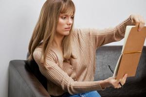 Confused sad blonde student lady examines drawings at copybook sitting in sofa couch at modern living room interior. Female client have shrink appointment in psychological help office. Copy space photo