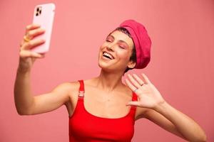 A young woman blogger with colored pink hair and a short haircut takes a picture of herself on the phone and broadcasts a smile in stylish clothes and a hat on a pink background monochrome style photo