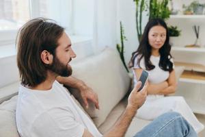 A man looks at his phone screen and smiles during an argument with his girlfriend. The angry and hurt woman looks in his direction and is sad. Family discord at home, phone addiction photo