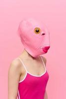 gracioso loco mujer en un rosado antecedentes en pie en un pescado cabeza máscara en un rosado fondo, conceptual Víspera de Todos los Santos disfraz Arte foto