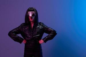 Shocked awesome brunet woman in leather jacket trendy specular sunglasses open mouth posing isolated with hands on waist in blue violet color light background. Neon party Cyberpunk concept. Copy space photo