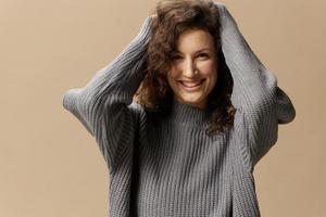 Cheerful smiling cute curly beautiful female in gray warm casual sweater touches head posing isolated on over beige pastel background. Fashion Sale offer. People emotions concept. Copy space photo