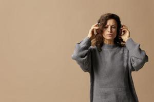 Pensive funny curly beautiful female in gray casual sweater headphones listen to her recording in studio posing isolated on over beige pastel background. Sound streaming platform concept. Copy space photo