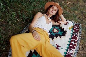 A woman in eco-friendly clothing in a hippie look is lying on a colored plaid smiling and looking at an autumn sunset in nature in the park. Lifestyle on a journey of harmony with the world photo