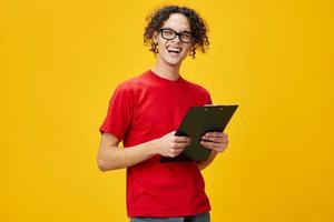 Emotional myopic young student man in red t-shirt funny eyewear holds tablet folder with study notes posing isolated on over yellow studio background. Free place for ad. Education College concept photo