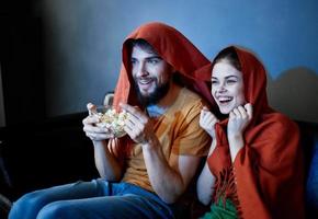 A man with a plate of popcorn and an emotional woman under a red blanket on the couch photo