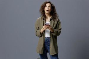 Thoughtful shocked pensive curly beautiful woman in casual khaki green shirt looks up got fake message using phone posing isolated on gray blue background. Social Media Influencer concept. Copy space photo