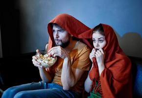 asustado mujer con un rojo tartán en su cabeza y un hombre con un plato de palomitas de maiz en un oscuro habitación foto
