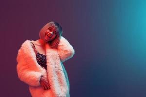 Happy adorable blonde woman in fluffy fur coat sparkly dress hold hand on hair posing isolated in blue turquoise red color light studio background. Neon party Fashion concept. Copy space Banner photo