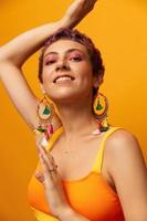 Portrait of a young woman with a short haircut and colored hair smiling and showing her tongue at the camera on an orange background with earrings accessories in the studio photo