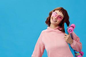 Happy cheerful pretty redhead lady in pink hoodie sunglasses with penny board posing isolated on blue studio background. Copy space Banner Offer. Fashion Cinema. Holiday activity concept photo