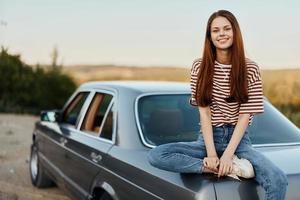 un joven mujer se sienta en el maletero de un coche y descansa después un difícil la carretera y admira naturaleza con un hermosa vista. parada es además parte de el viaje foto