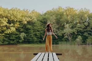 un joven niña en un hippie Mira soportes en un puente por un lago en el otoño y mira a el puesta de sol foto