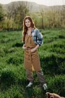 un granjero mujer en su trabajo ropa, tartán camisa y delantal, soportes en el campo en el verde césped y sonrisas en el ajuste Dom después un difícil días trabajo foto
