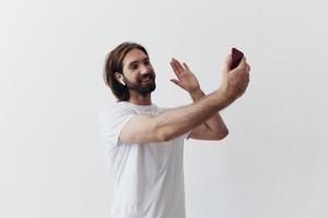 un masculino blogger grabando en video él mismo en su teléfono y chateando con personas en línea con un sonrisa en un blanco camiseta en contra un blanco pared foto