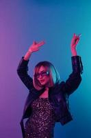 Enjoyed sexy beautiful blonde woman in leather jacket sparkly dress trendy sunglasses dance hold hands up show v-sign posing isolated in blue pink color light studio background. Neon party concept photo