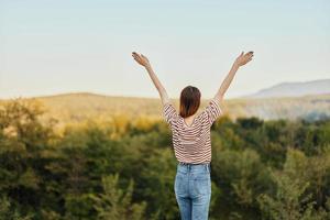 A young woman stands with her back to the camera with her hands up in a T-shirt and jeans in nature and enjoys a beautiful view of the mountains. Autumn travel to nature lifestyle photo