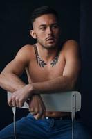 Modelling snapshots. Pensive serious tanned attractive handsome naked man sit on chair smoking looks aside posing isolated in black studio background. Fashion offer. Copy space for ad. Closeup photo