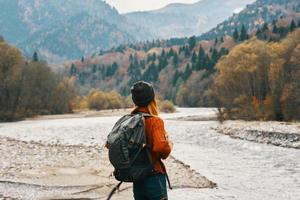 woman in sweater with a backpack rest in the mountains near the river in autumn in nature and tall trees model photo