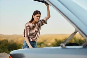 el joven mujer conductor mira a el coche en horror por apertura el capucha. un coche rompió abajo en un la carretera viaje foto