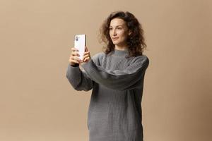 Joyful curly beautiful female in gray casual sweater doing selfie with phone posing isolated on over beige pastel background. Social media, network, distance communication concept. Copy space photo