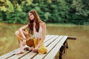 A young hippie woman sits on a lake bridge wearing stylish eco clothes and smiling photo
