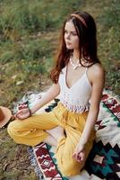 A young hippie woman meditates in nature in the park, sitting in a lotus position on her colorful plaid and enjoying harmony with the world in eco-clothing photo