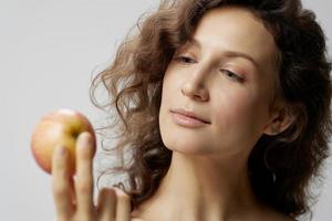 Smiling pensive cute curly beautiful woman in basic white t-shirt looks at apple enjoy healthy food posing isolated on over white background. Natural Eco-friendly products concept. Copy space photo