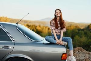 A woman car driver sits on the trunk of a car and smiles admiring a beautiful view of autumn nature and mountains photo