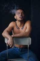 Modelling snapshots. Pensive serious tanned attractive handsome naked man sit on chair smoking looks aside posing isolated in black studio background. Fashion offer. Copy space for ad. Closeup photo