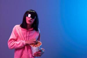 Shocked awesome brunet woman in pink hoodie trendy specular sunglasses eat popcorn open mouth posing isolated in blue violet color light background. Neon party Cinema concept. Copy space photo