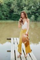 A young woman in a hippie look and eco-dress dancing outdoors by the lake wearing a hat and yellow pants in the summer sunset photo