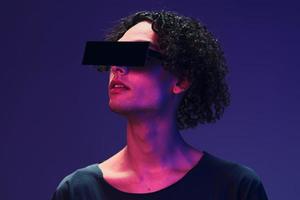 Interested stylish tanned curly man in black t-shirt trendy eyewear posing isolated on color pink blue studio background. Cool fashion offer. Virtual Reality New Collection concept. Copy space for ad photo