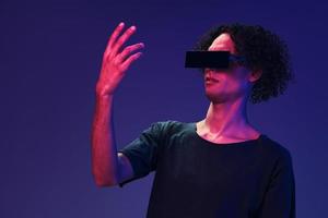 Excited stylish tanned curly man in black t-shirt eyewear holds invisible object posing isolated on color pink blue background. Cool fashion offer. Virtual Reality New Collection concept. Copy space photo