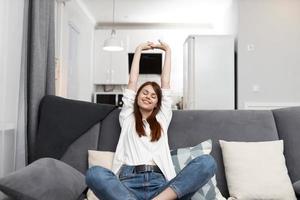 alegre mujer extensión sí misma mientras sentado en el sofá descansando Departamento interior foto