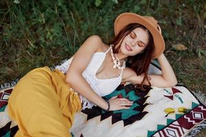 A woman in eco-friendly clothing in a hippie look is lying on a colored plaid smiling and looking at an autumn sunset in nature in the park. Lifestyle on a journey of harmony with the world photo