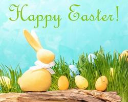 Yellow Easter bunny on textured wood, decorative eggs in green grass on turquoise Words Happy Easter photo