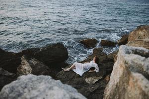 atractivo mujer con largo pelo en un blanco vestir acostado en un Roca en un blanco vestir inalterado foto