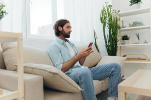 A freelance man in a white T-shirt, blue jeans, and shirt sits on the couch with his phone at home on his day off and plays games photo