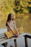 un joven contento mujer con un hippie sonrisa se sienta en el lago apuntalar en un puente y medita en un loto actitud vistiendo eco ropa hecho de natural materiales en armonía con naturaleza en el otoño foto