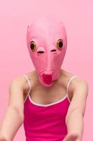 A woman wearing a silicone Halloween mask in the shape of a pink fish with big yellow eyes looks at the camera against a pink background photo