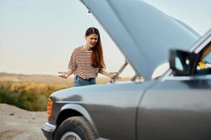 un triste mujer tiene abrió el capucha de un roto abajo coche y es mirando para el porque de el Descompostura en un la carretera viaje solo con su manos untado aparte y gritos en ira foto