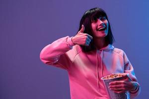 Cheerful awesome brunet woman in pink hoodie with popcorn show thumb up posing isolated in blue violet color light studio background. Neon party Cyberpunk Cinema concept. Copy space photo