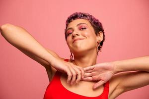 Fashion portrait of a woman with a short haircut of purple color and a smile with teeth in a red top on a pink background dancing happily photo