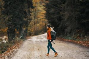 woman in jeans jacket with a backpack walks along the road in the autumn forest photo