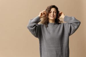 Relaxing happy smiling curly beautiful female in gray casual sweater headphones enjoy fav playlist posing isolated on over beige pastel background. Sound streaming platform concept. Copy space photo