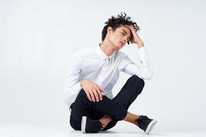 un chico con Rizado pelo en un blanco camisa, zapatillas y pantalones se sienta en el piso en un brillante habitación foto