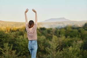 A young woman stands with her back to the camera with her hands up in a T-shirt and jeans in nature and enjoys a beautiful view of the mountains. Autumn travel to nature lifestyle photo