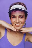 retrato de un deportivo Moda mujer posando sonriente con dientes a el cámara con elegante pendientes en su orejas en un púrpura yoga chandal y un transparente gorra en un púrpura monocromo antecedentes foto